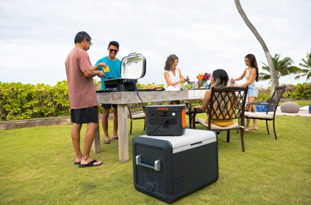  Un groupe d'amis et de famille fait un barbecue dans un jardin, avec un appareil Jackery et une glacière.