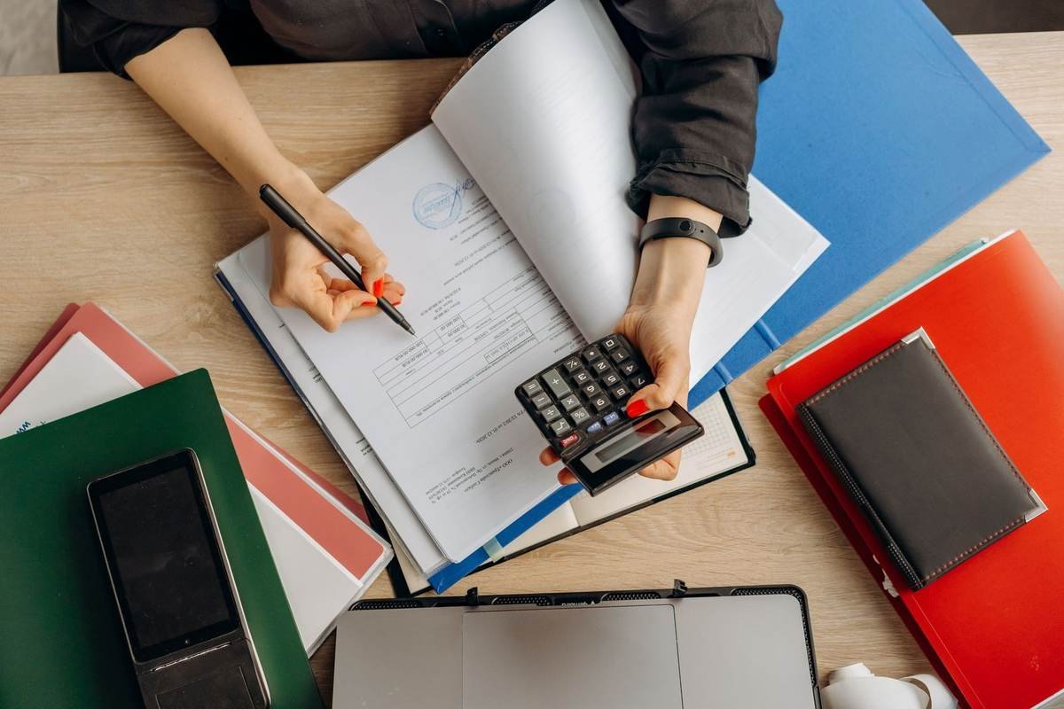 Person écrivant dans un livre avec une calculatrice, classeurs et téléphone sur une table Person écrivant dans un livre avec une calculatrice, classeurs et téléphone sur une table