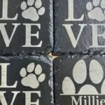 Gros plan de quatre sous-verres carrés en ardoise, dont trois affichent le mot « LOVE » avec des empreintes de pattes pour la lettre « O » et un quatrième affichant une empreinte de patte avec le mot « Millie ». Les sous-verres sont disposés en grille sur une surface en bois.