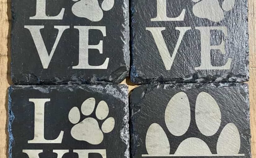 Gros plan de quatre sous-verres carrés en ardoise, dont trois affichent le mot « LOVE » avec des empreintes de pattes pour la lettre « O » et un quatrième affichant une empreinte de patte avec le mot « Millie ». Les sous-verres sont disposés en grille sur une surface en bois.